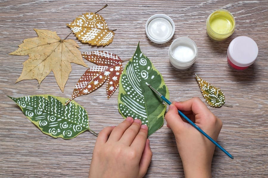 Girl paints patterns leaf. Gouache, brush and various autumn leaves on a wooden table. Children's art project. Colorful Hand-painted on dry autumn leaves